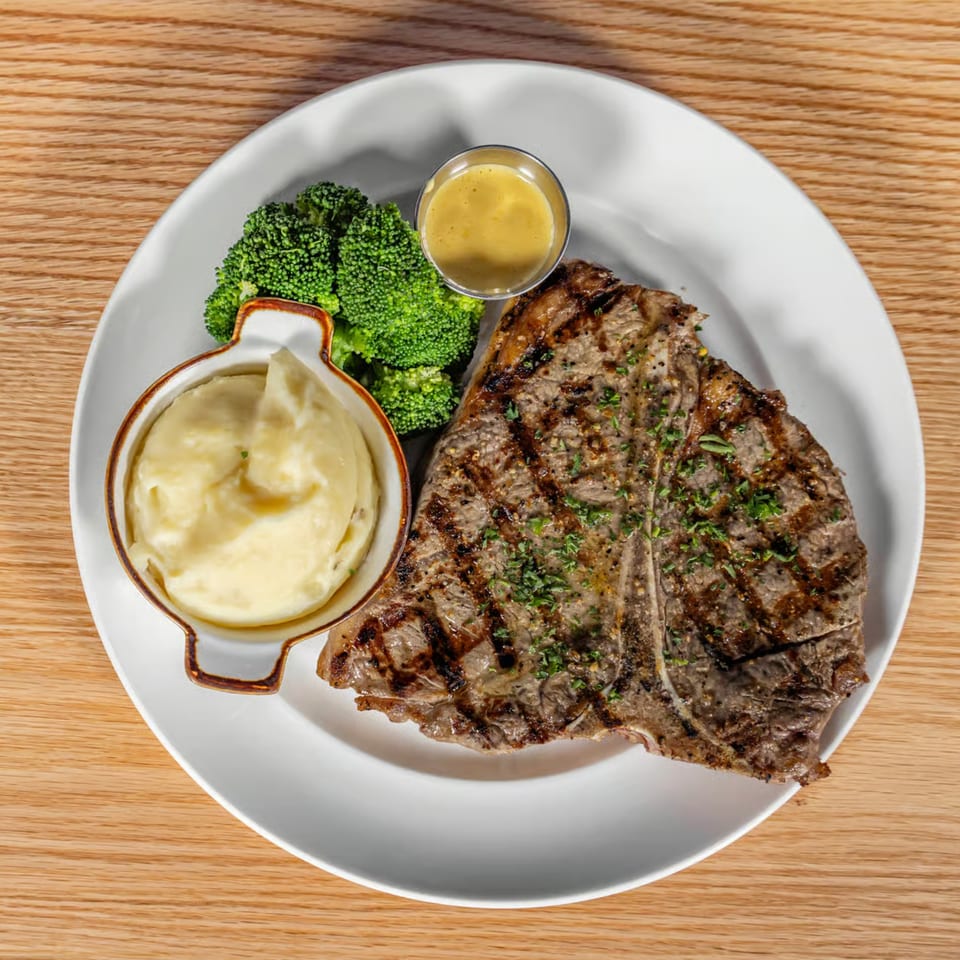 Grilled T-bone steak with mashed potatoes, broccoli, and a side of mustard sauce on a white plate.