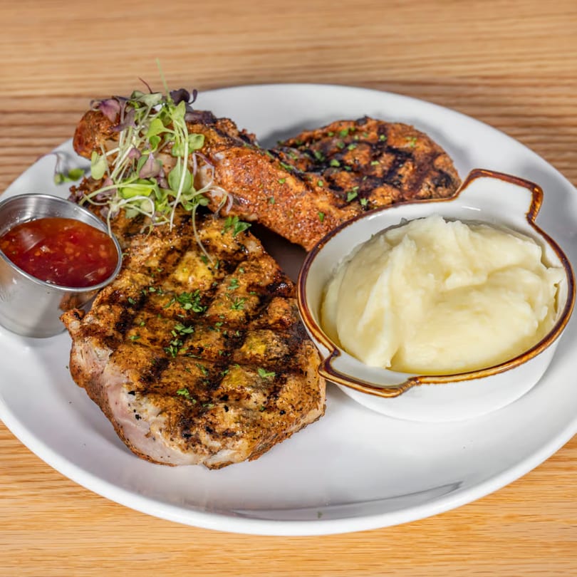 Grilled pork chops with mashed potatoes and a side of sweet chili sauce on a white plate.