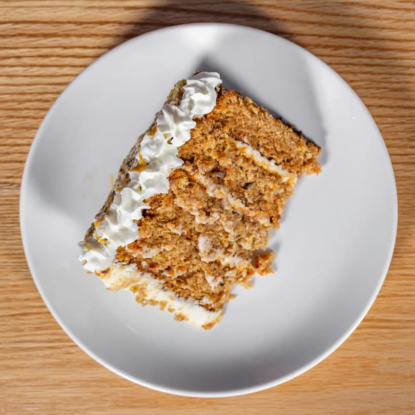 Slice of carrot cake with white frosting and whipped cream on a white plate.