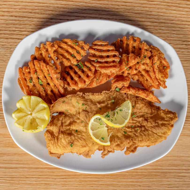 Crispy fried fish fillet with waffle fries and lemon wedges on a white plate.