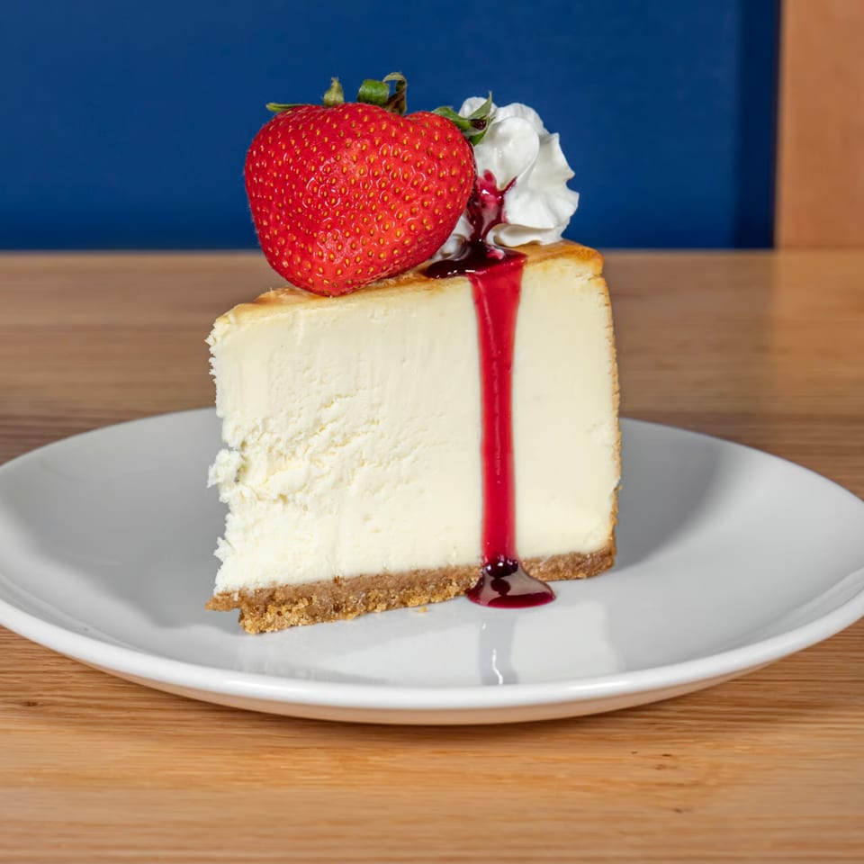 Slice of cheesecake with a graham cracker crust, topped with a strawberry, whipped cream, and berry sauce.
