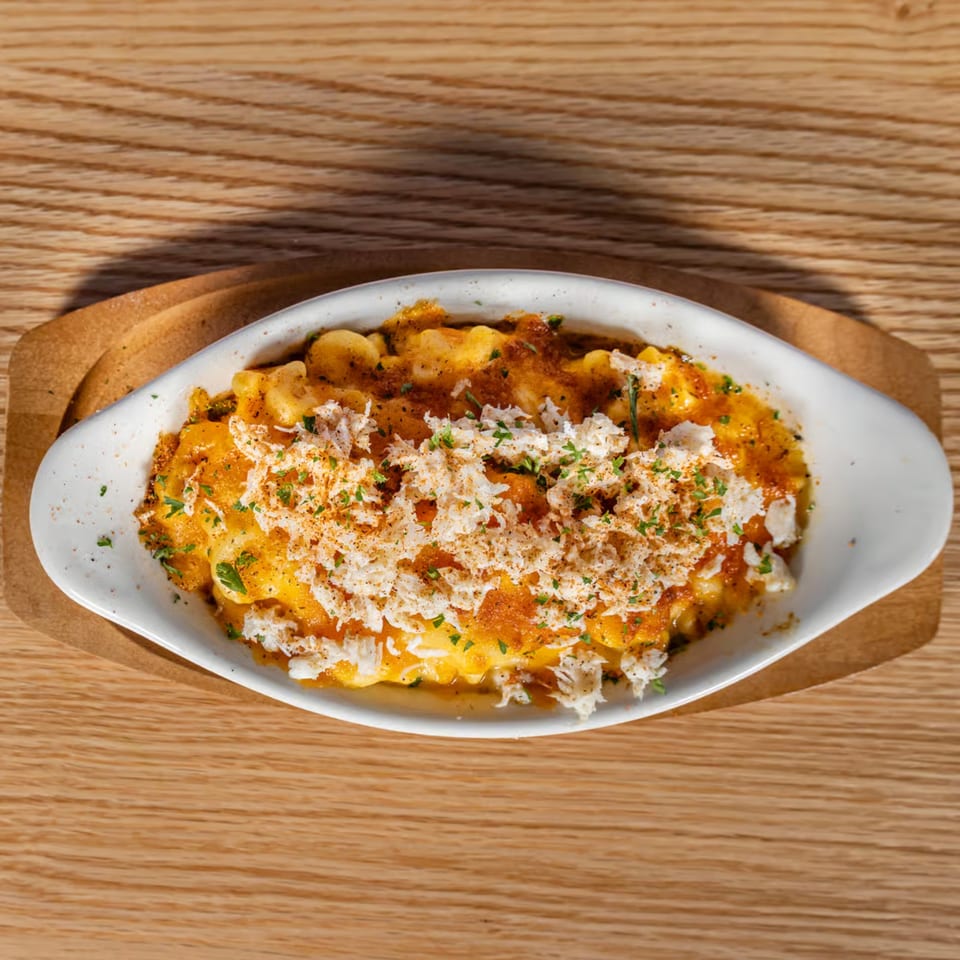 A white oval dish of golden mac and cheese topped with crab meat and spices on a wooden table.