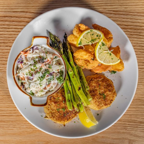 A white plate with fried shrimp, crab cakes, asparagus, coleslaw, and lemon slices.