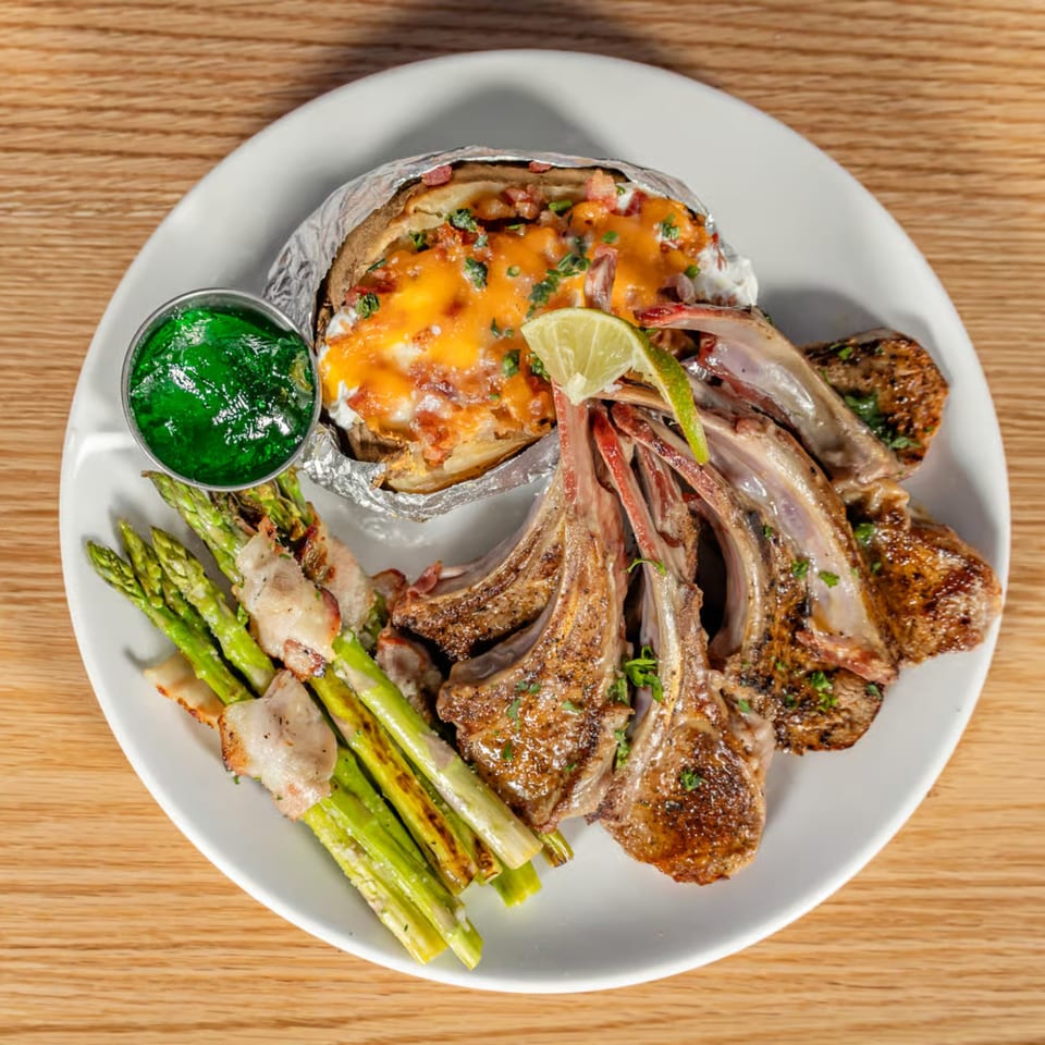 Grilled lamb chops, bacon-wrapped asparagus, and a loaded baked potato with lime on a white plate.