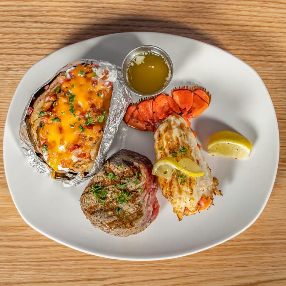 Surf and turf meal with steak, lobster tail, baked potato, and melted butter on a white plate.
