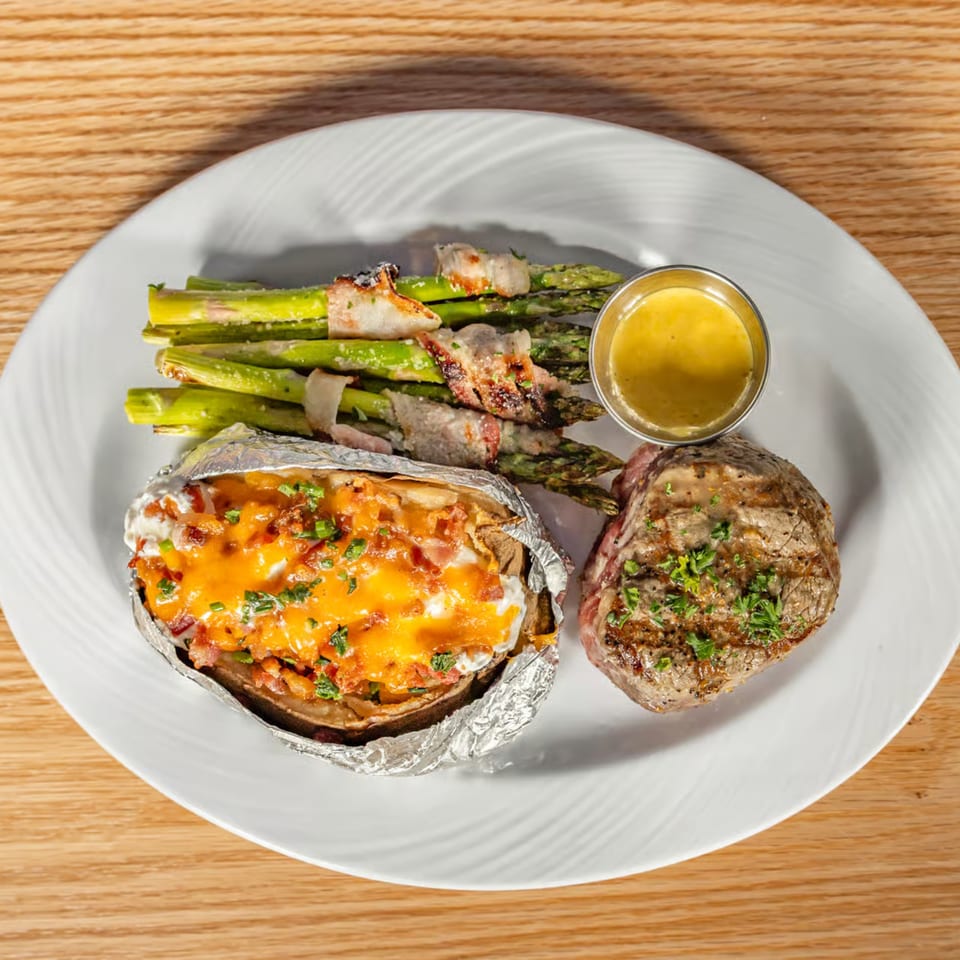 Grilled steak, bacon-wrapped asparagus, and a loaded baked potato with cheese and chives on a white plate.