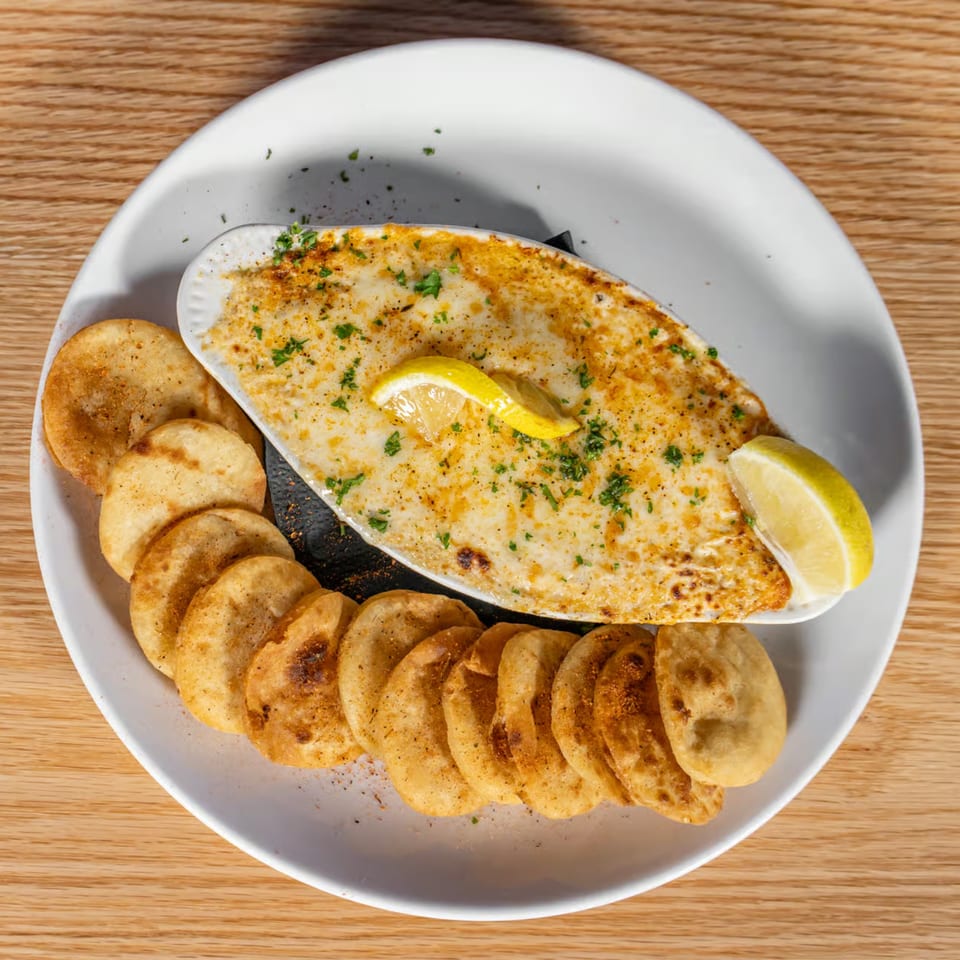 Baked dip with melted cheese, herbs, and lemon wedges, served with crispy round chips on a white plate.