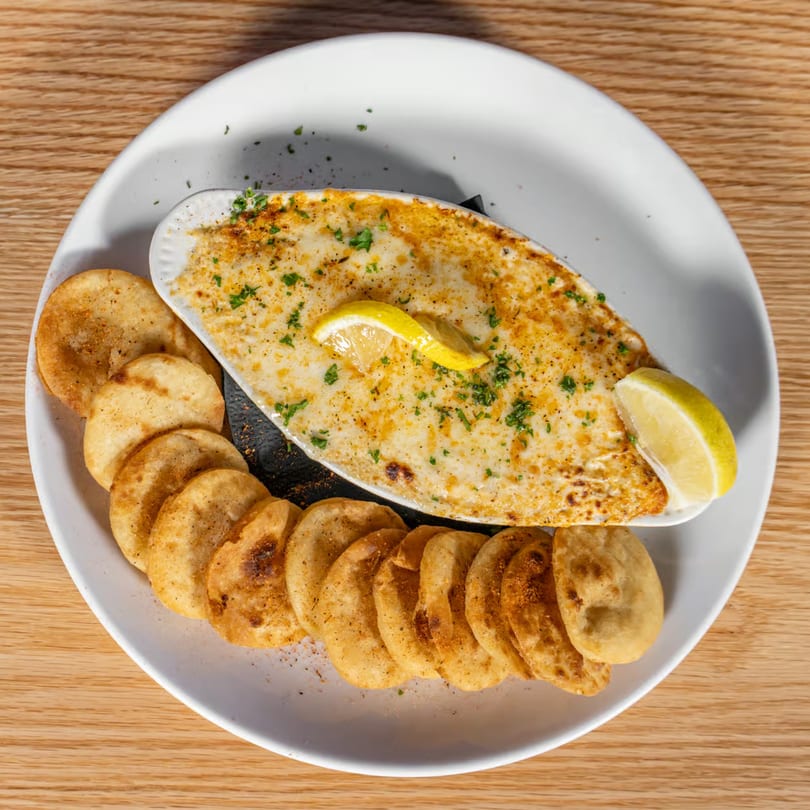 Baked dip with melted cheese, herbs, and lemon wedges, served with crispy round chips on a white plate.