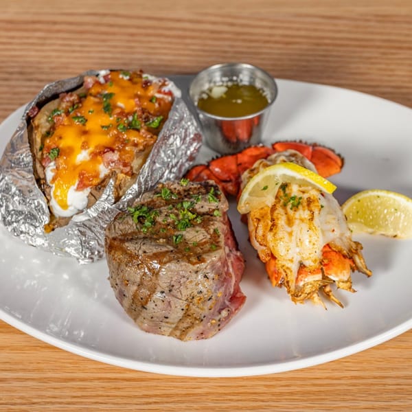 Steak, lobster tail with lemon, loaded baked potato, and melted butter on a white plate.